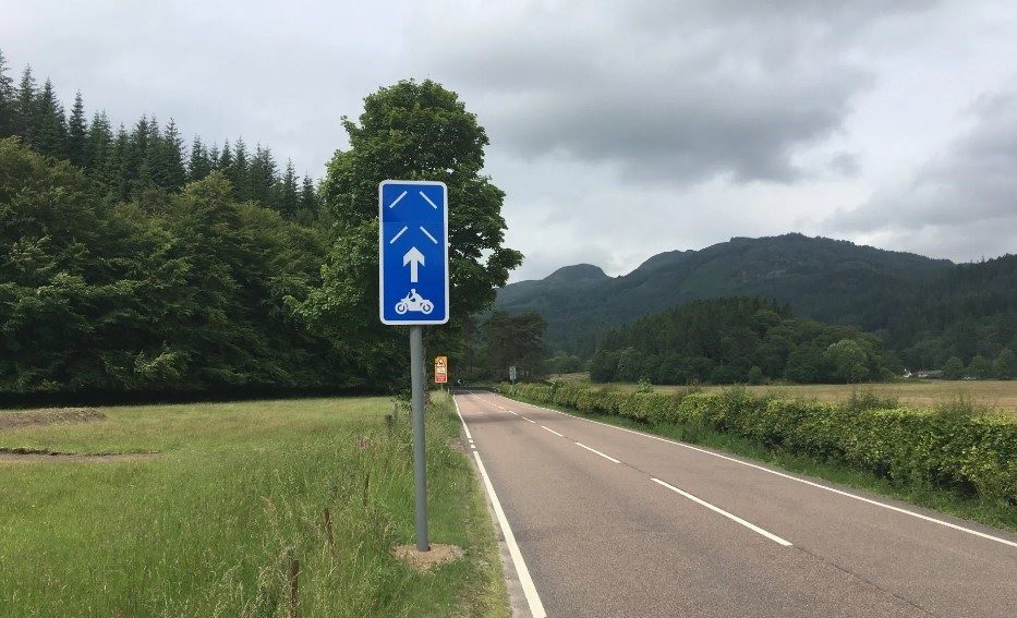 Road in Scottish countryside, on the side of the road is a blue sign that is targeted to motorbike riders as part of Project PRIME